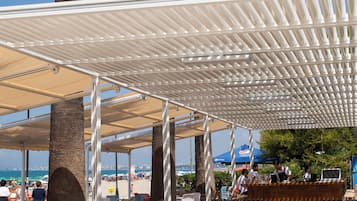 An outdoor seating area with wicker chairs and a covered section, palm trees, and a view of the beach.