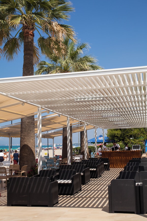 An outdoor seating area with wicker chairs and a covered section, palm trees, and a view of the beach. - Premium Beach Hotel (Golem)