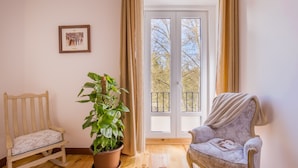 Una habitación con piso de madera, una mecedora, un sillón acolchado, una planta en maceta y una puerta corrediza de vidrio que da a un balcón al aire libre.