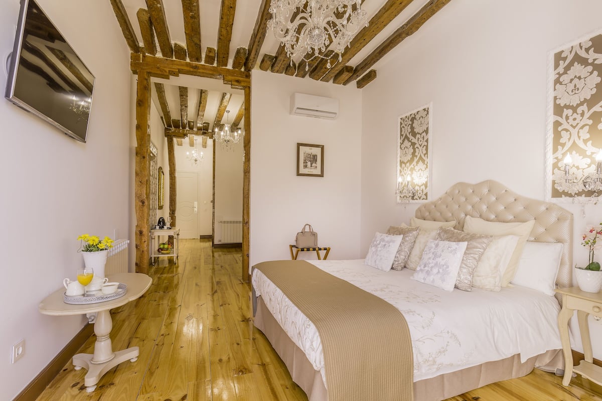 A bedroom with a large bed, wooden beams, a chandelier, a bedside table, and a wall-mounted TV.