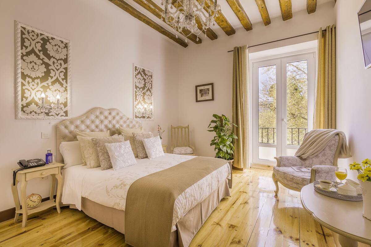 A bedroom with a large bed, a bedside table with a lamp, a chair, and a view of the outdoors through a window.