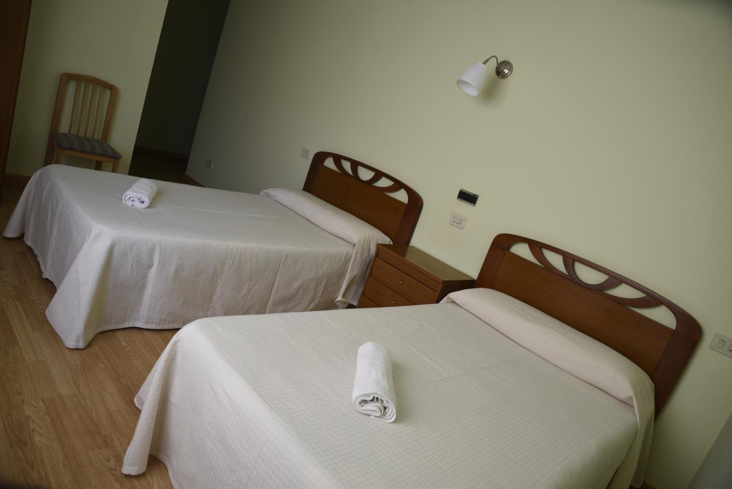 Two single beds with wooden headboards, a chair, and a wall-mounted light fixture in a room with a plain wall and wooden flooring.
