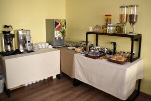 Montaje de bufé con estación de café, dispensador de fruta y diversos alimentos sobre mesas.
