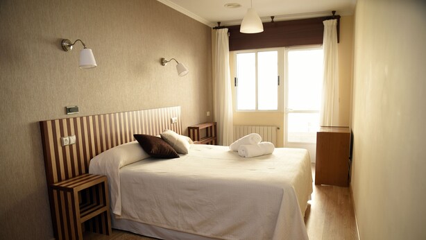 A neatly arranged hotel room with a large bed, bedside tables, a nightstand, a radiator, and a window with curtains. - Hotel Villa De Ribadeo (Ribadeo)