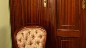 A wooden chair with a cushioned seat and backrest, positioned in front of a wooden door with a brass handle. - Hotel Villa De Ribadeo (Ribadeo)