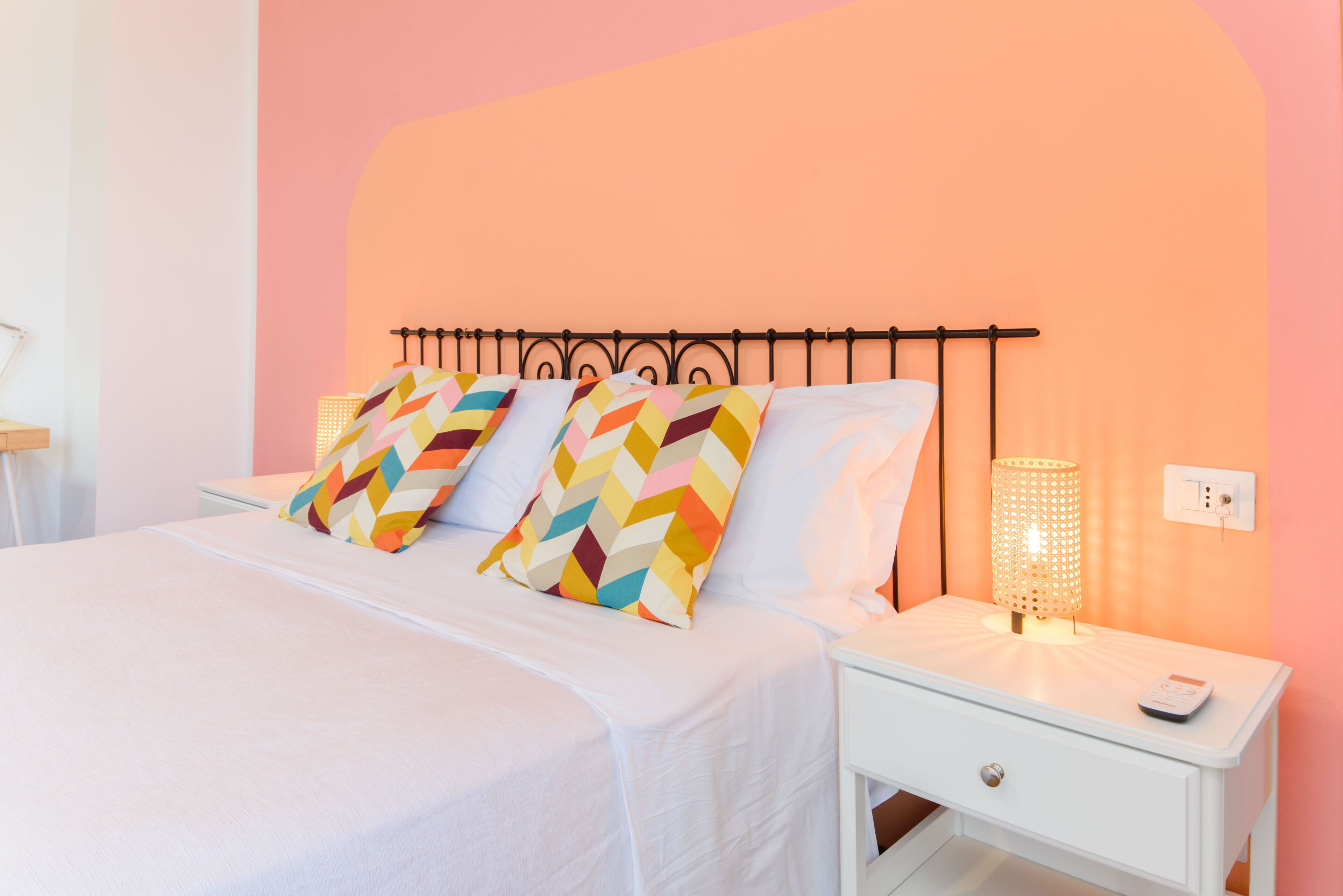 Una cama bien hecha con almohadas coloridas, una mesita de noche con lámpara y un enchufe en la pared.
