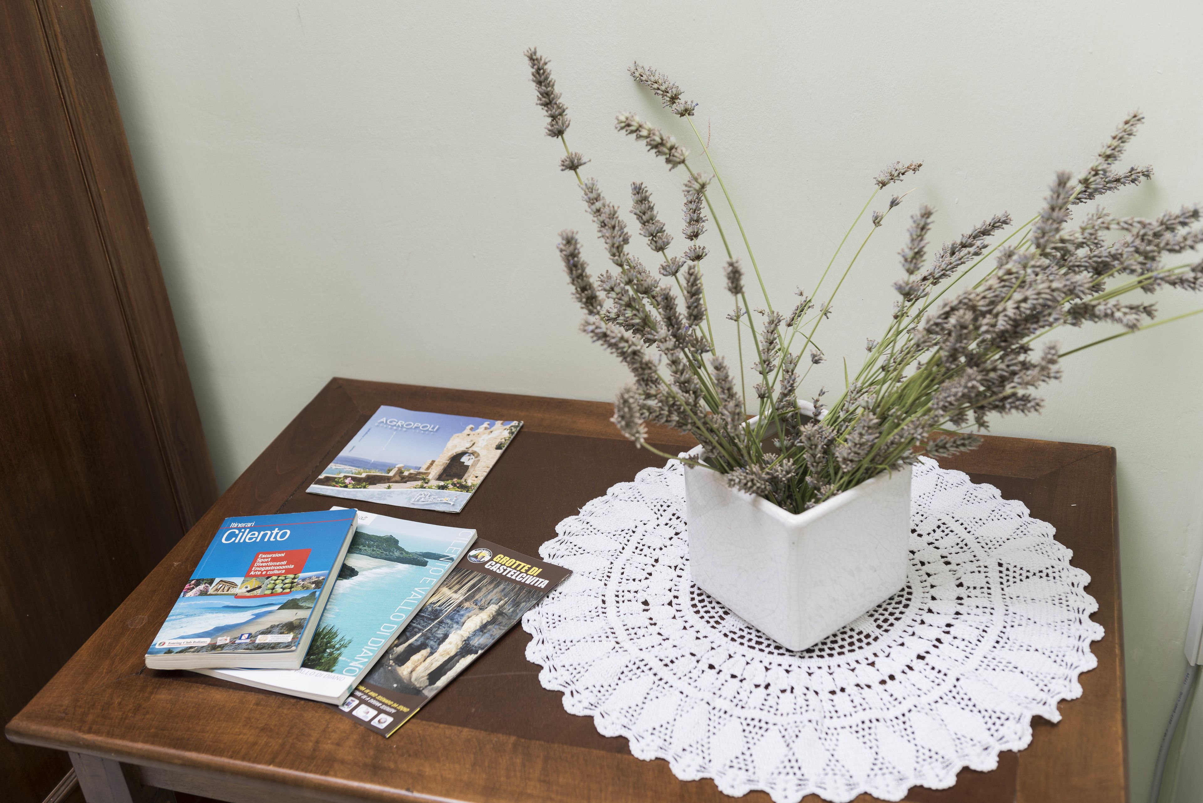Una mesa de madera con un mantel individual blanco, un jarrón blanco con lavanda y folletos de viajes.