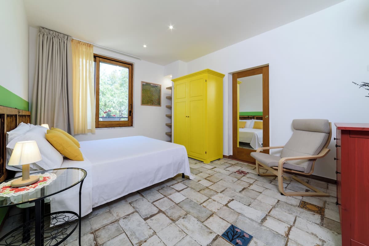 A bedroom with a bed, a chair, a wardrobe, a table with a lamp, and a window with curtains.