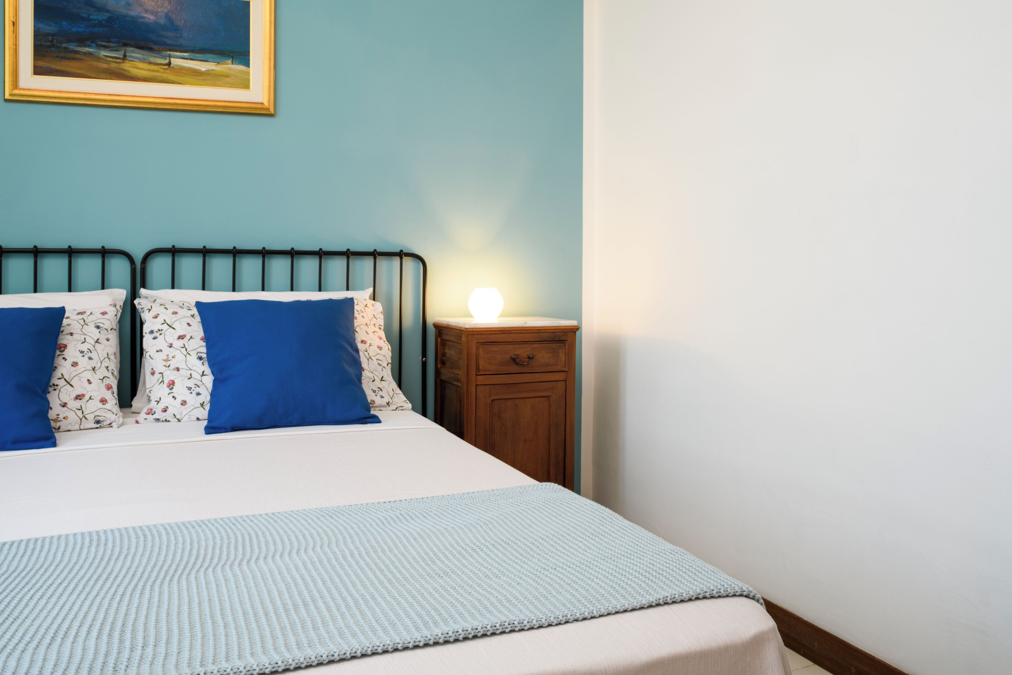 A neatly made bed with blue and floral pillows, a bedside table with a lamp, and a painting on the wall.