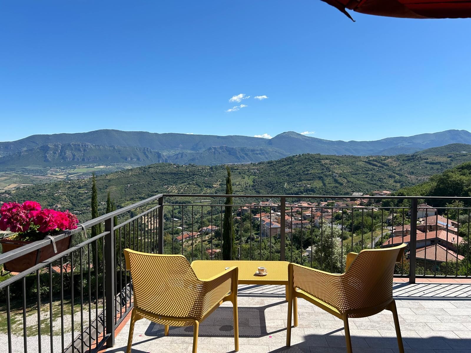 Un balcon avec des chaises jaunes et une table, d’où l’on peut admirer un paysage montagneux avec un village en contrebas.