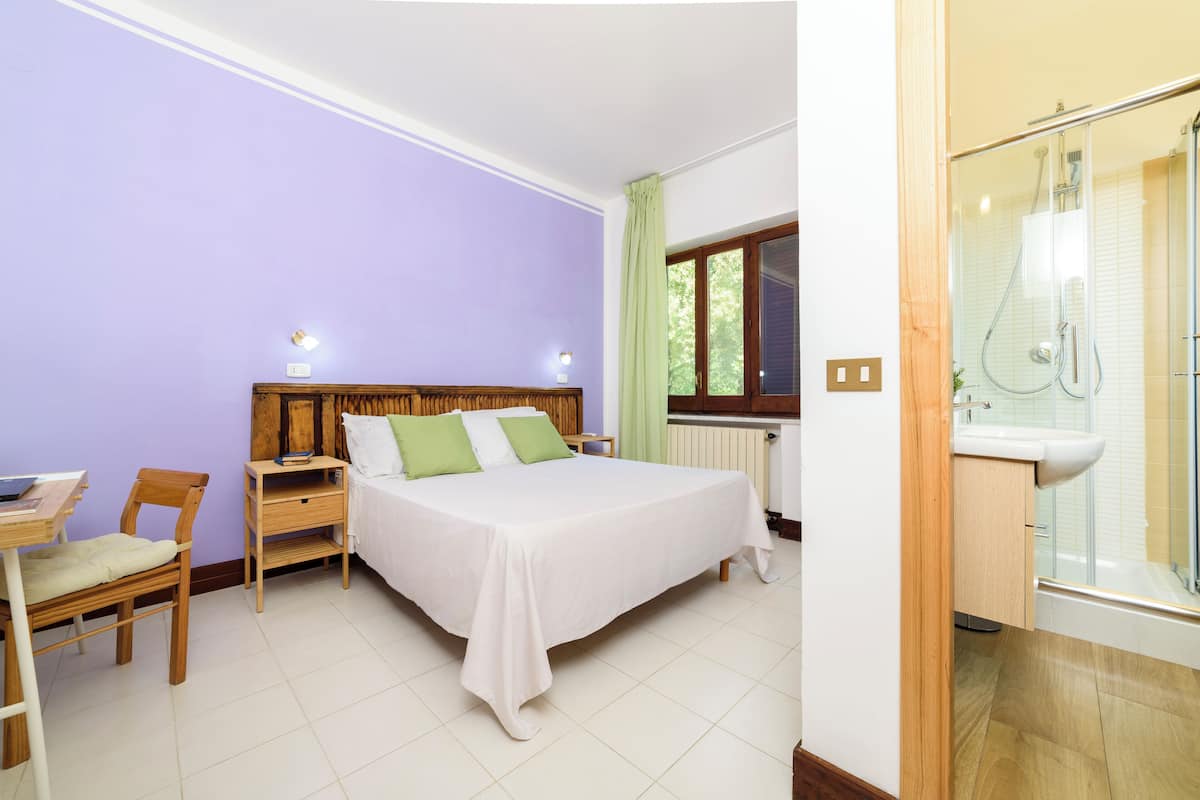 A bedroom with a bed, bedside tables, a chair, a window, and a bathroom with a shower and sink.