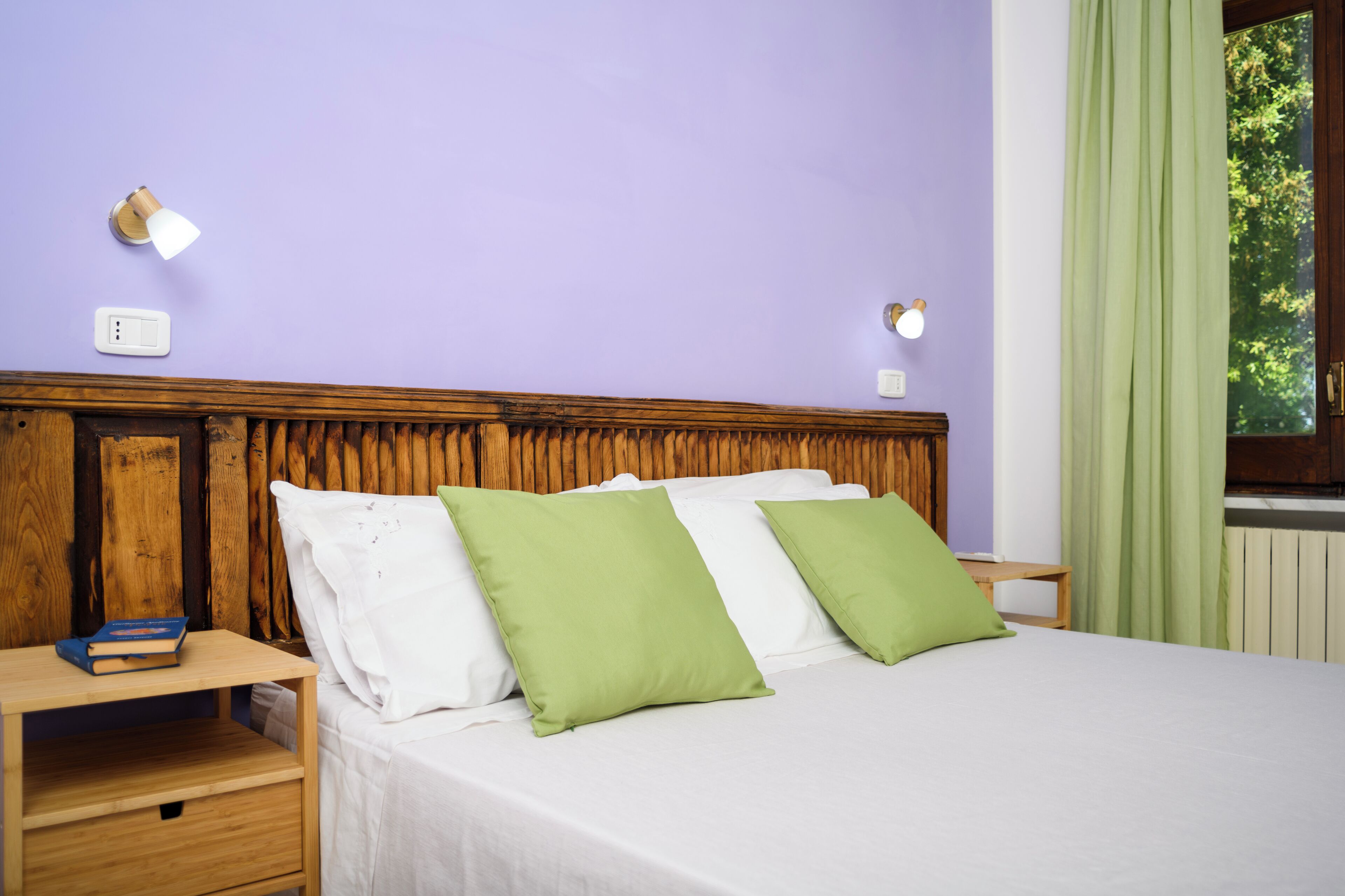 A bedroom with a wooden headboard, two bedside tables, a bed with white and green pillows, a nightstand with a book, and a window with green curtains.