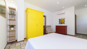 Un dormitorio con cama, un armario amarillo, cómoda de madera, una silla y una estantería con plantas.
