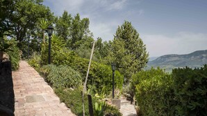 A brick pathway leading through a lush garden with various plants and trees, a lamp post, and a view of distant hills. - Albachiara Casa Di Campagna B&B (Ogliastro Cilento)