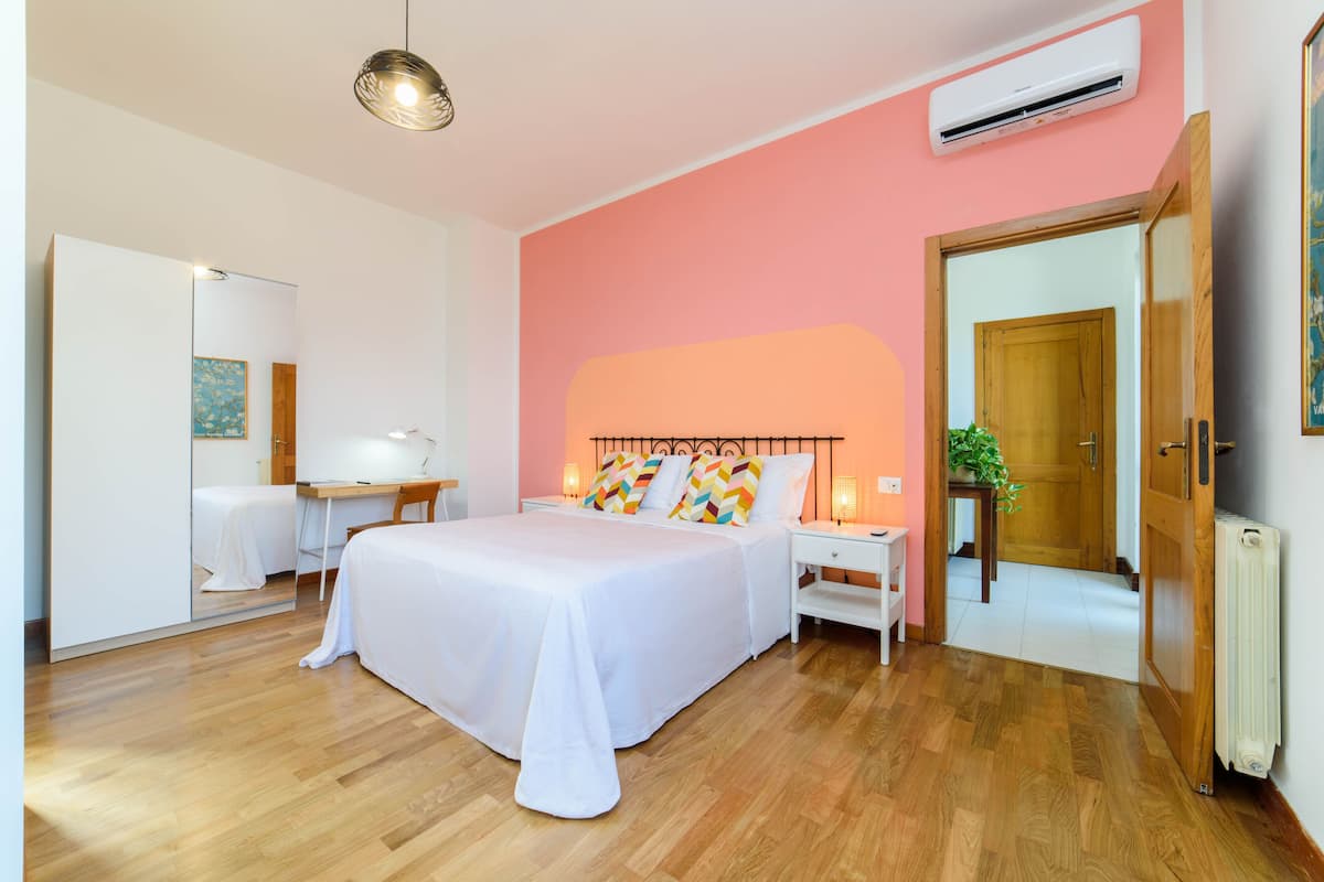 A bedroom with a large bed, bedside tables, a desk, and a chair. There is a door leading to another room and an air conditioning unit on the wall.