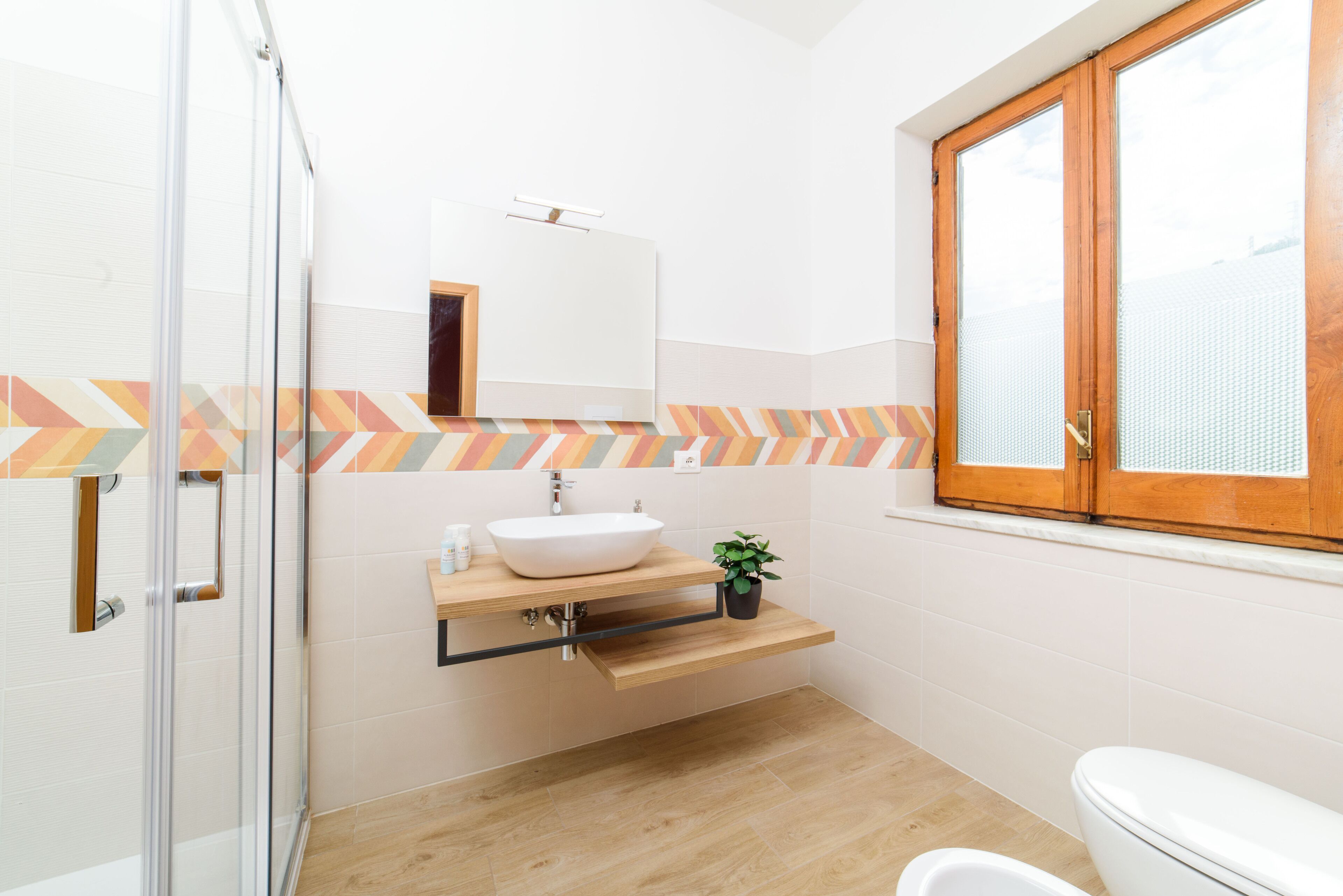 Un bagno moderno con cabina doccia in vetro, lavandino bianco con piano in legno e finestra in legno con parete a motivo geometrico.