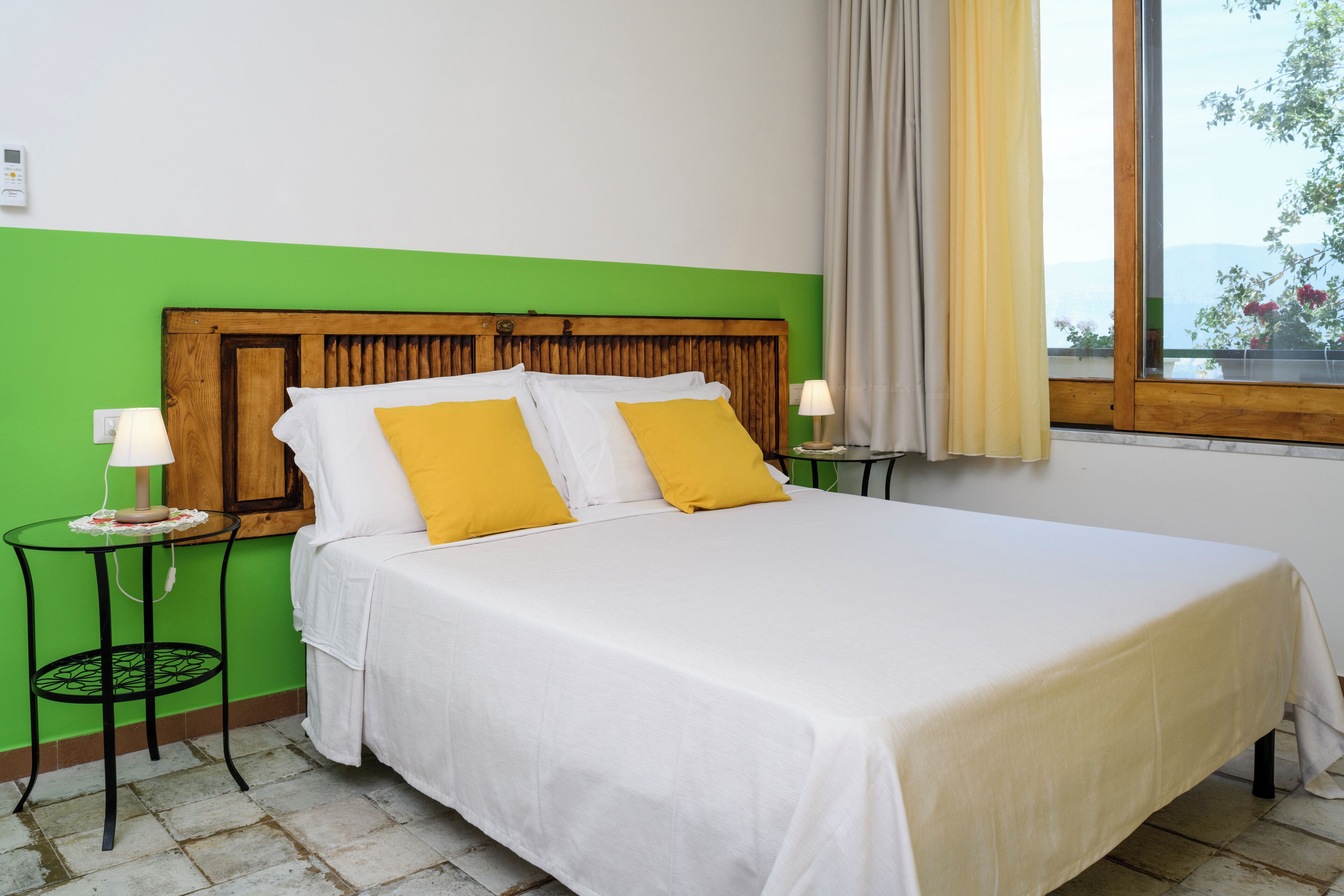 A bed with white bedding and two yellow pillows, a wooden headboard, a small table with a lamp, and a window with a view of greenery and mountains.