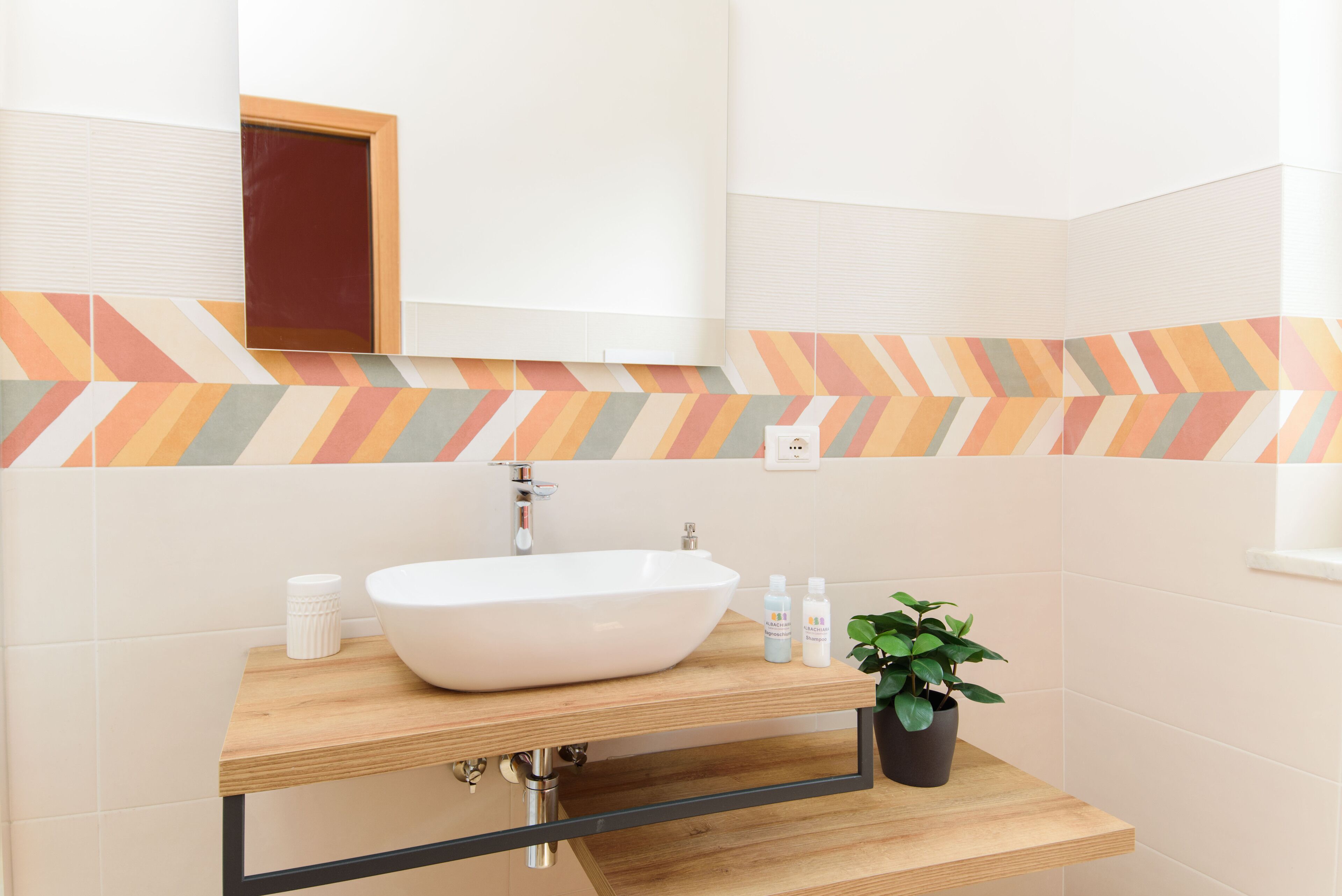 Un baño moderno con un lavamanos blanco, encimera de madera y un espejo encima. Hay una planta en maceta sobre la encimera y un pequeño florero blanco.