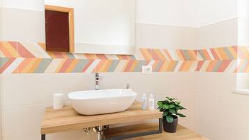 Un baño moderno con un lavamanos blanco, encimera de madera y un espejo encima. Hay una planta en maceta sobre la encimera y un pequeño florero blanco.