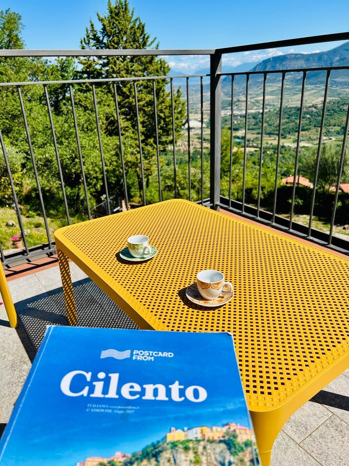 Une table en métal jaune sur laquelle reposent deux tasses, une carte postale intitulée « Carte postale du Cilento », et une vue sur un paysage montagneux, visible à travers une rambarde en métal.