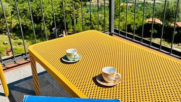 A yellow metal table with two cups on it, a postcard titled "Postcard from Cilento," and a view of a mountainous landscape through a metal railing.