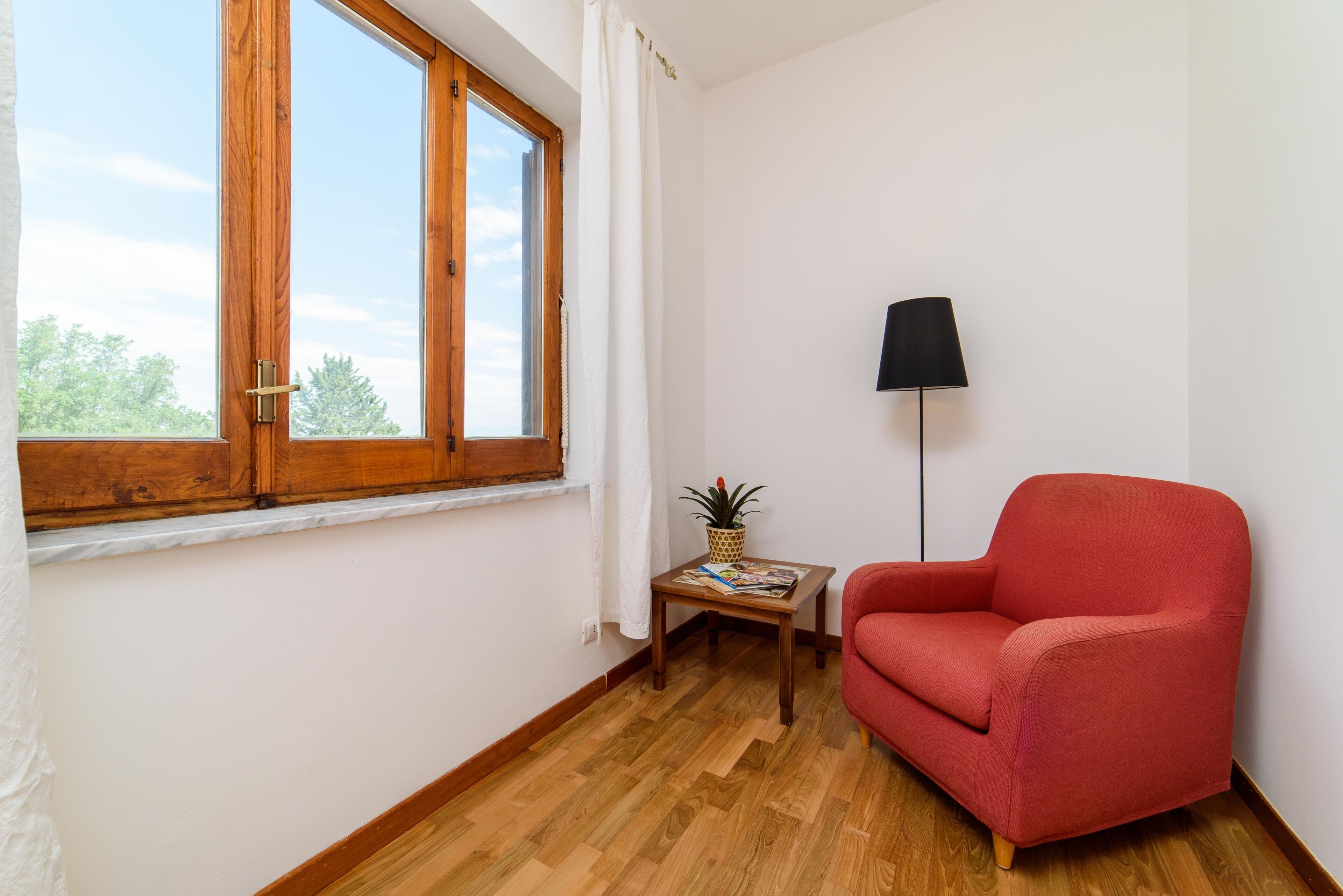 A room with a wooden floor, a red armchair, a small table with a plant, and a floor lamp.