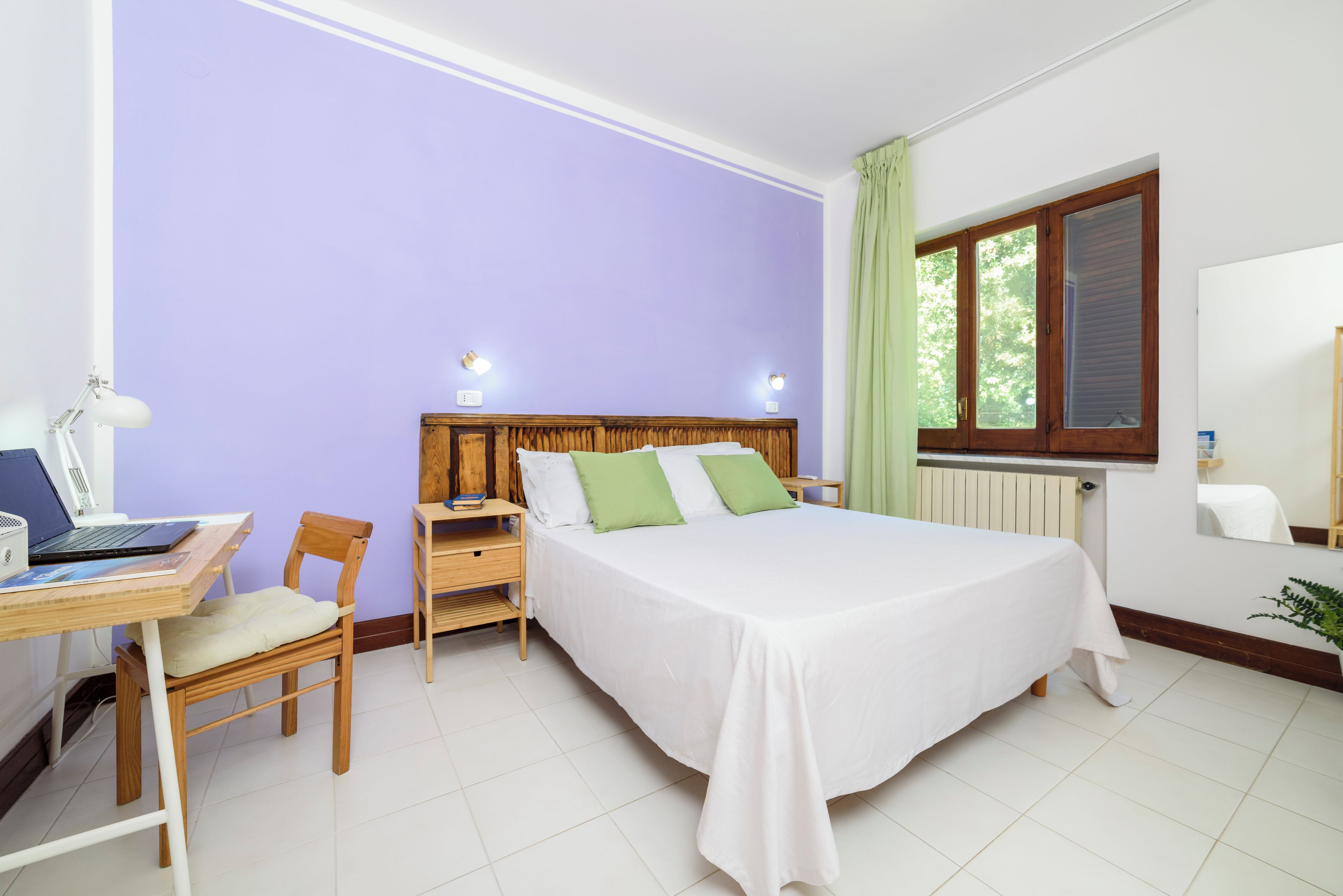 A bedroom with a bed, a desk with a laptop, a chair, and a window with curtains.