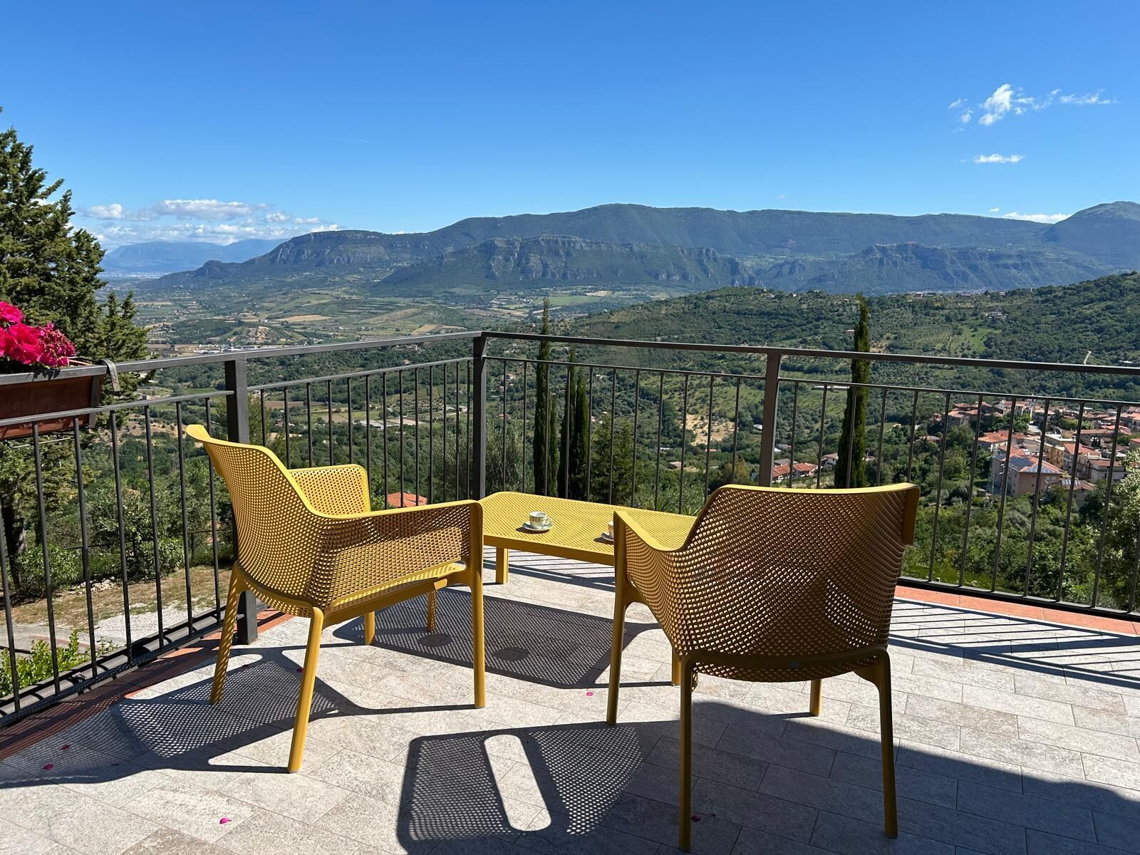 Un balcón con sillas de mimbre amarillo y una pequeña mesa, con vistas a un paisaje montañoso.