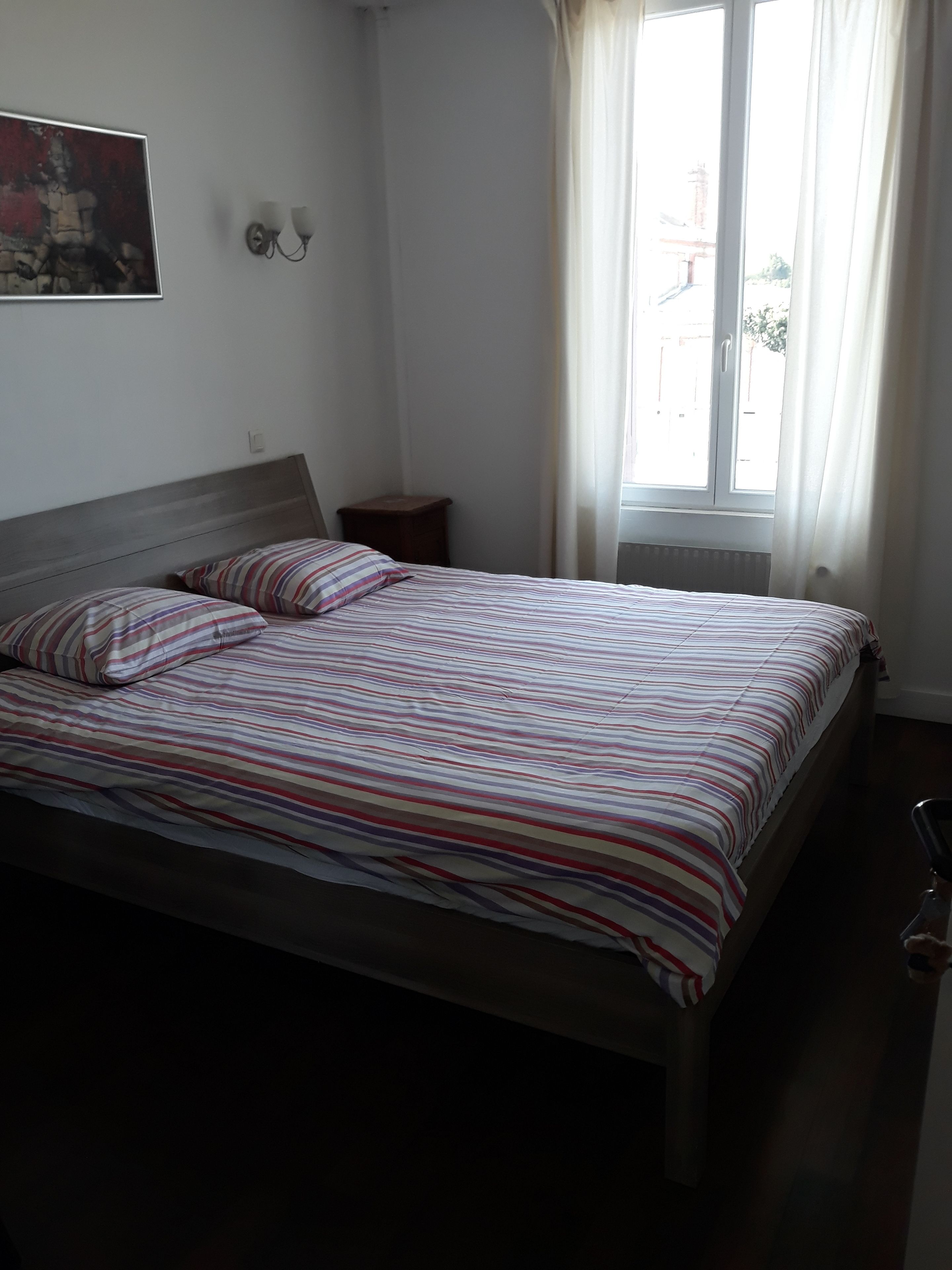 A bedroom with a bed, a nightstand, a wall-mounted lamp, and a window with curtains.