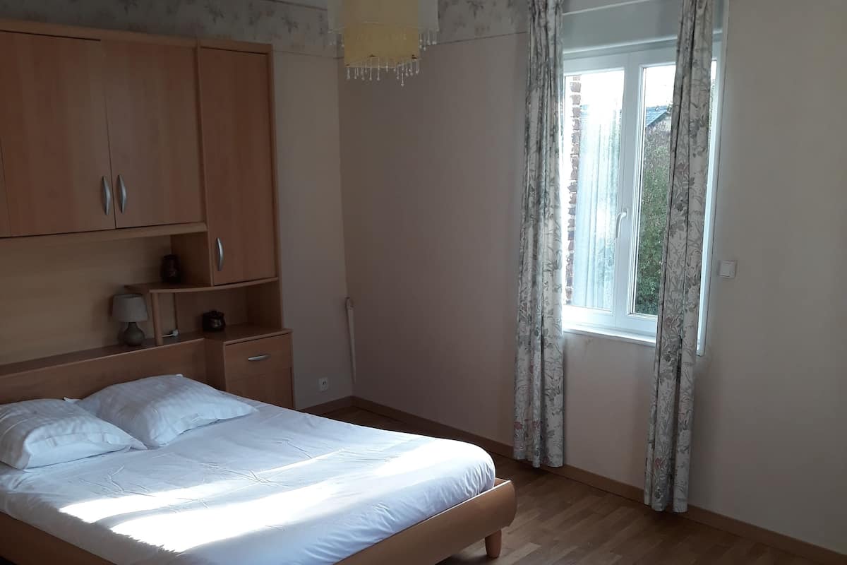 A bedroom with a wooden bed, a hanging light fixture, a window with curtains, and a wardrobe.