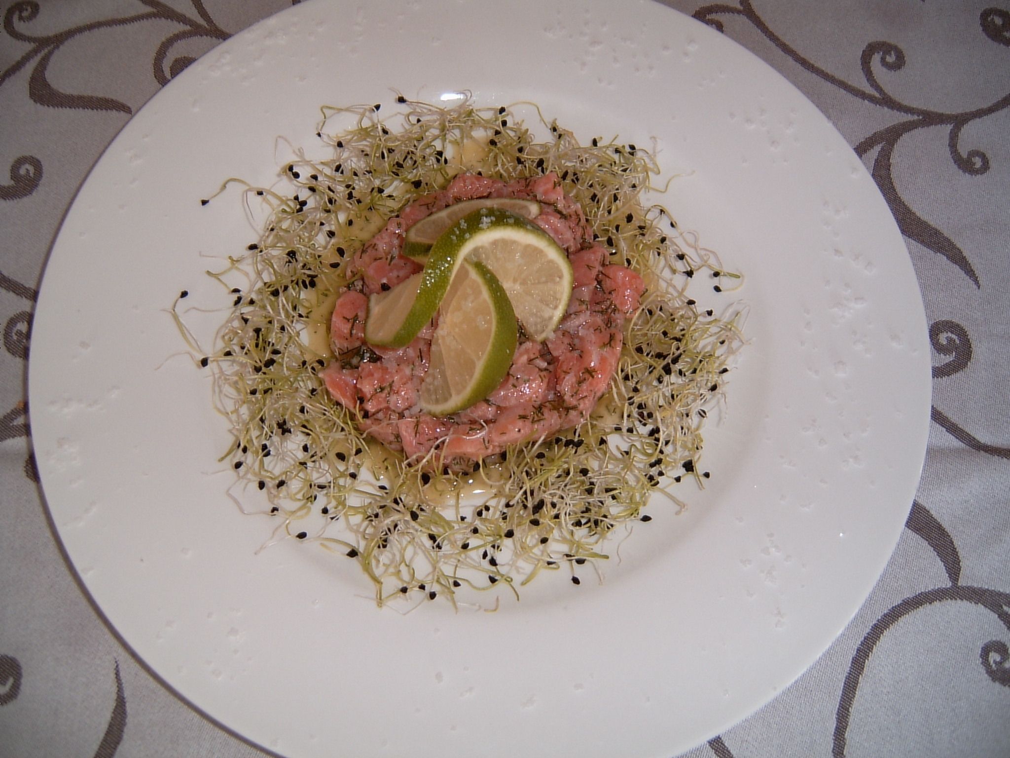 Une assiette blanche contenant une portion de viande rosée, des pousses et des tranches de citron vert.