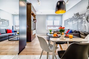Un salon moderne avec un coin repas, agrémenté d’une fresque représentant un paysage urbain, d’une table à manger avec des chaises et d’un canapé avec des coussins colorés.