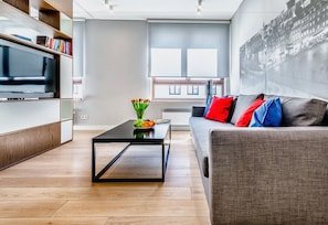 A modern living room with a flat-screen TV, a coffee table with a vase of flowers, a sofa with colorful cushions, and a large window with a cityscape view. - Business Center Phoenix Apartments (Warsaw)