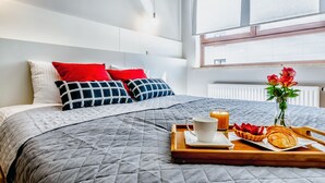 A neatly made bed with a grey quilt, red and blue pillows, and a tray with breakfast items including a croissant, strawberries, and a cup of coffee. - Business Center Phoenix Apartments (Warsaw)