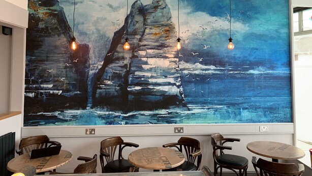 A large abstract painting of a coastal scene with three hanging light bulbs. - Carrick Rooms (Port St Mary)