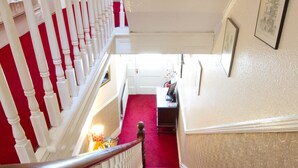 A grand staircase with red carpeted steps and white balustrades, leading to a landing area with a desk and framed artwork on the wall. - Carrick Rooms (Port St Mary)