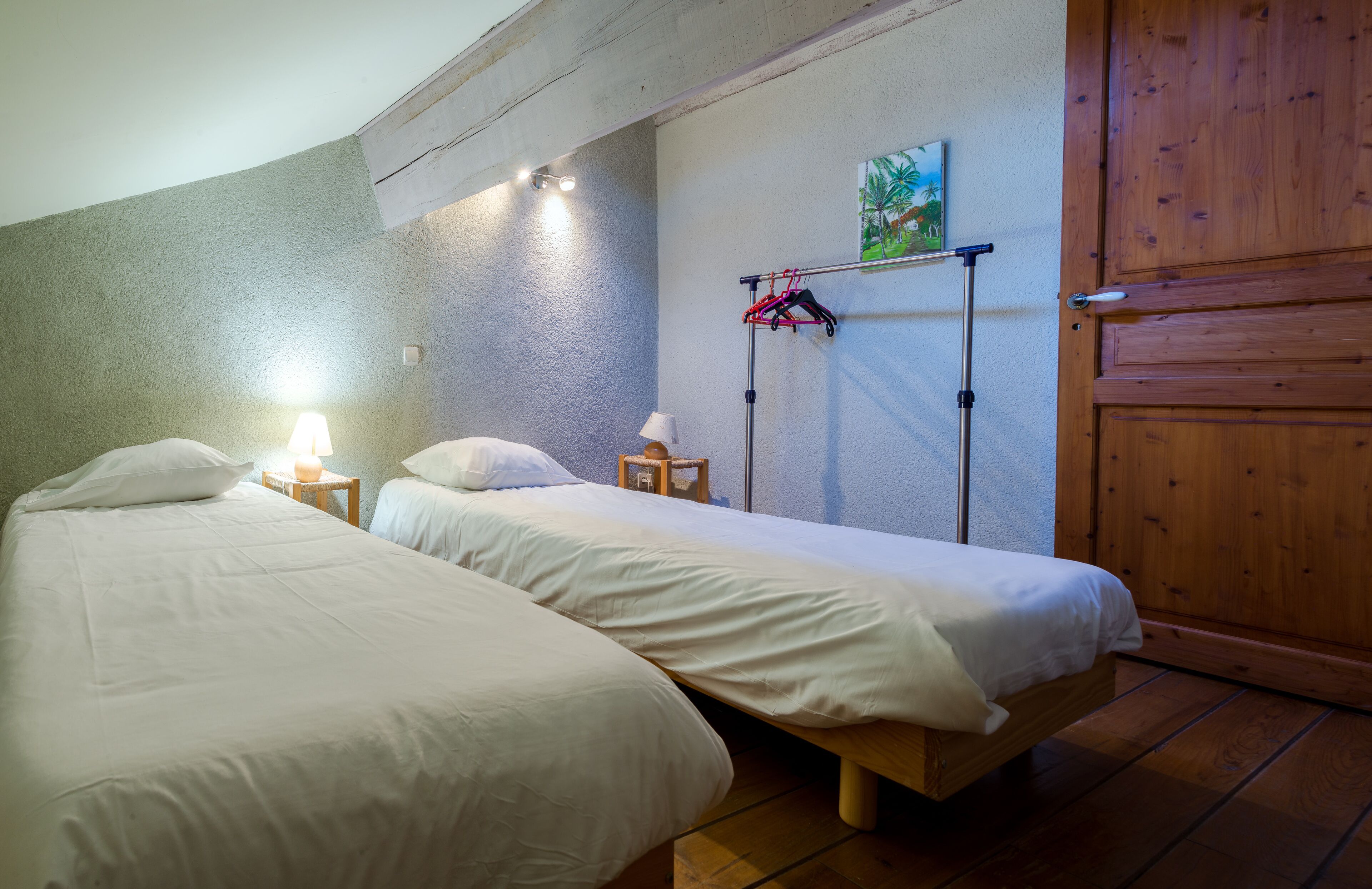 A room with two single beds, a wooden door, a wall-mounted drying rack, and a small table with a lamp.