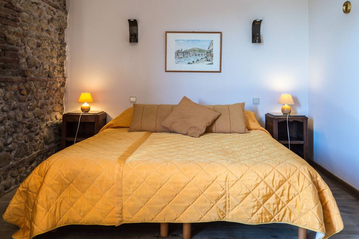 A bed with a quilted duvet, two bedside tables with lamps, a framed picture on the wall, and a stone wall to the left.