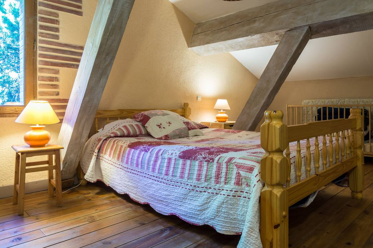 A cozy bedroom with a wooden bed, a nightstand with a lamp, and a window with a view of trees.