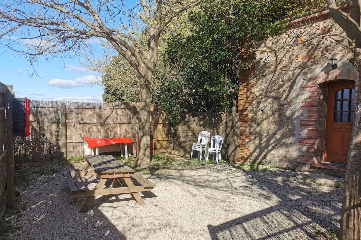 A rustic outdoor area with a wooden table and chairs, a red tablecloth, and a brick building with a window.