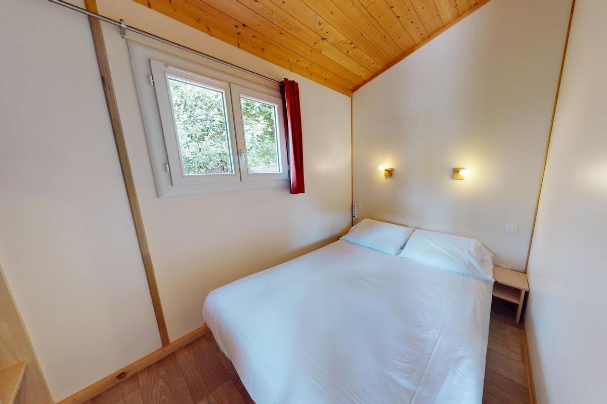 A single bed in a room with wooden ceiling and walls, a window with curtains, and two wall-mounted lights.