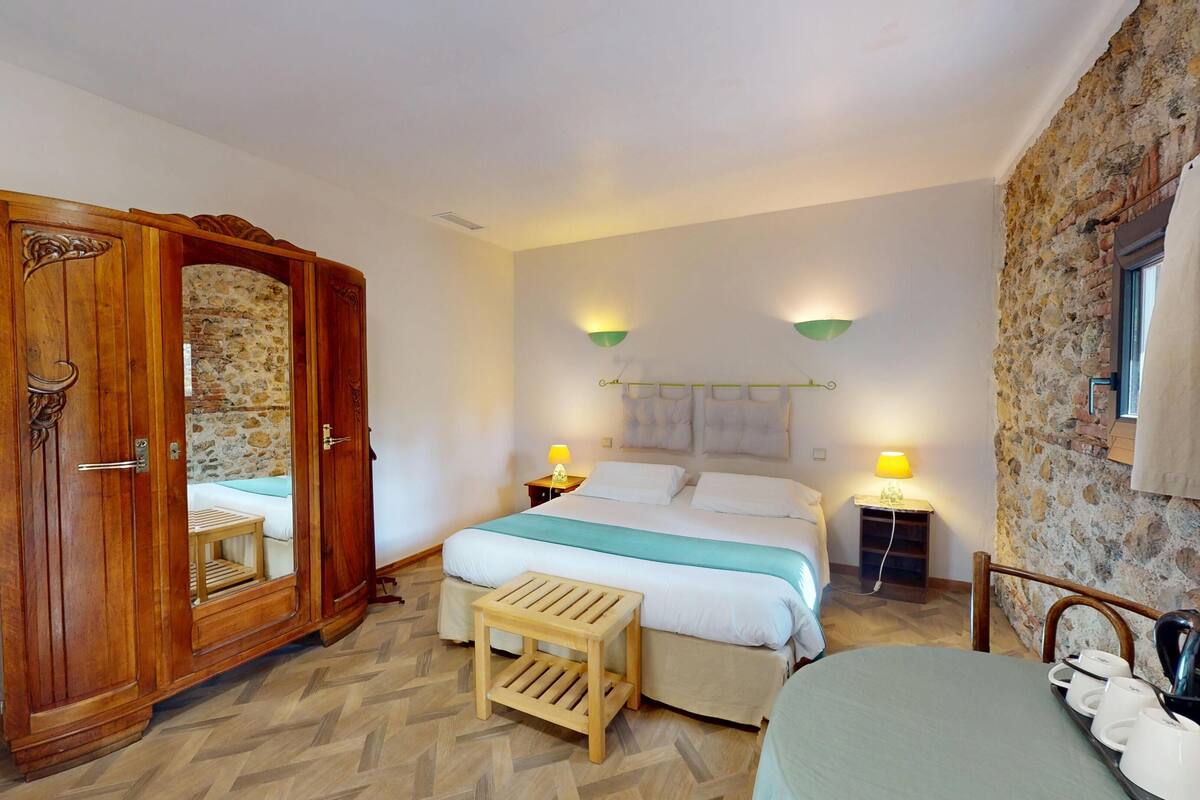 A bedroom with a wooden wardrobe, a bed with white bedding, two bedside lamps, and a stone wall.