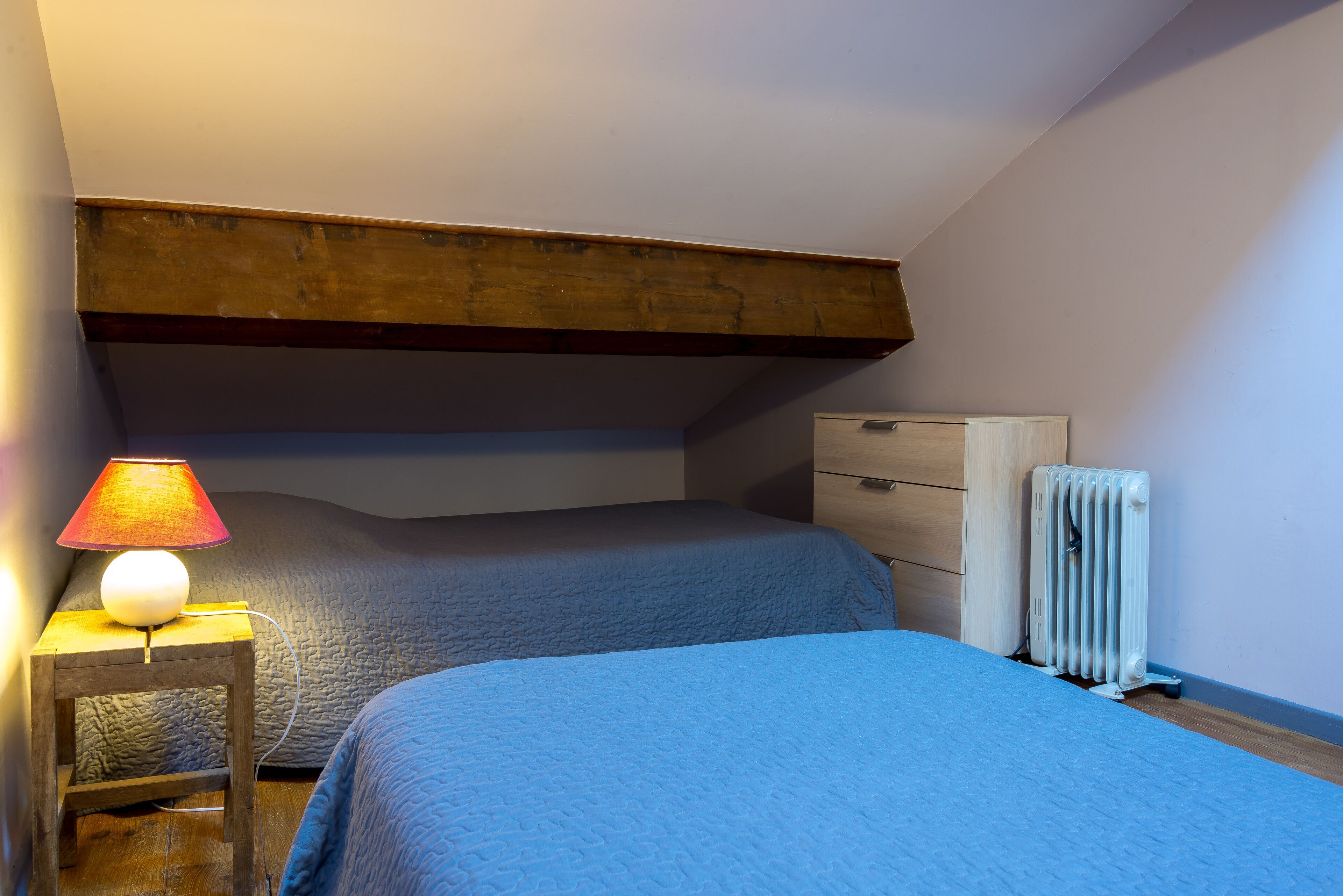 Une chambre avec un plafond en pente, une table de chevet avec une lampe, un radiateur et un lit recouvert d’une couverture bleue.