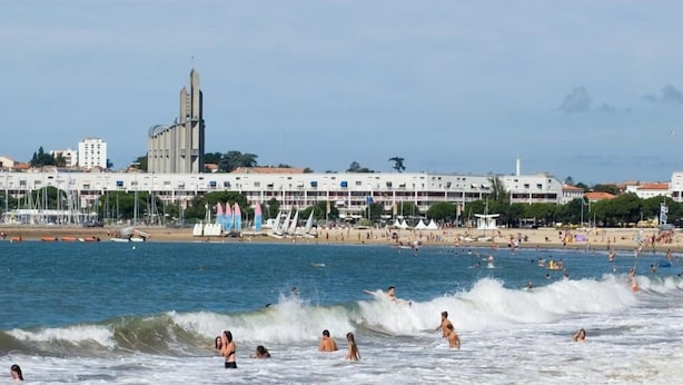 A beach with people swimming and surfing, a row of white buildings, and sailboats in the background. - Le Crystal Hôtel (Royan)