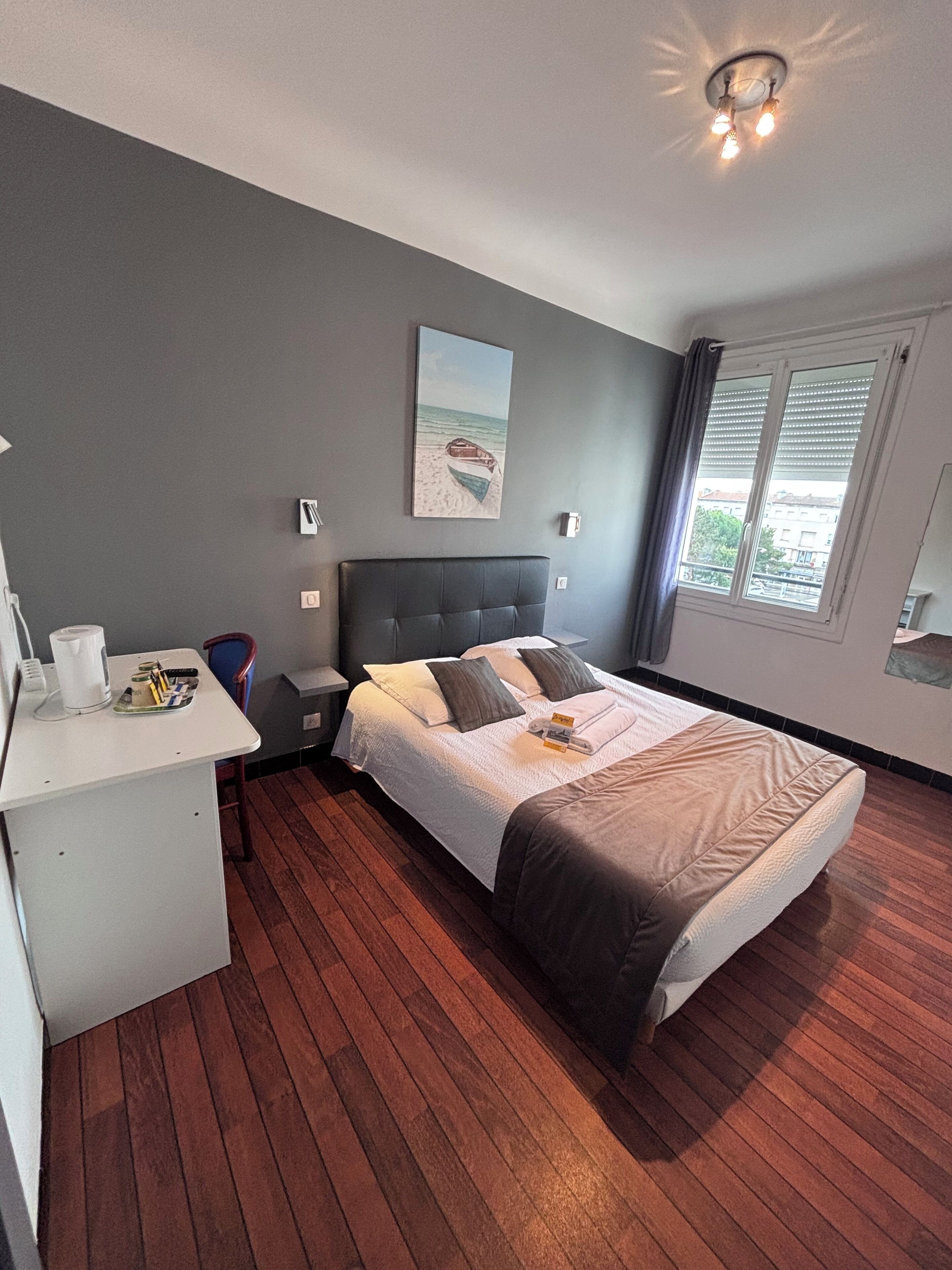 A bedroom with a bed, a desk, a chair, a painting, and a window.