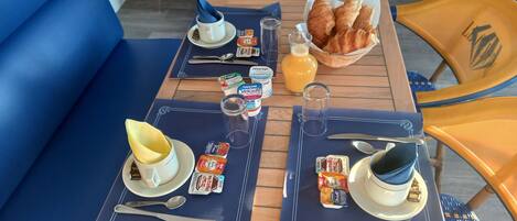 A breakfast setup with two tables, each with a blue placemat, a cup, a plate with a pastry, a glass of orange juice, and a bottle of Danone yogurt.