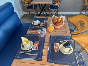 A breakfast setup with two tables, each with a blue placemat, a cup, a plate with a pastry, a glass of orange juice, and a bottle of Danone yogurt. - Le Crystal Hôtel (Royan)