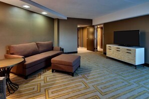 A modern hotel room with a sofa, ottoman, TV stand, and a patterned carpet. - Mount Vernon Hotel and Event Center (Mount Vernon)
