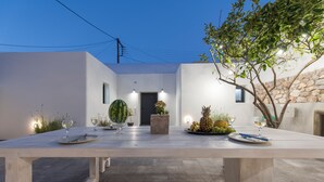 A modern outdoor dining area with a long table set for two, surrounded by plants and a stone wall.