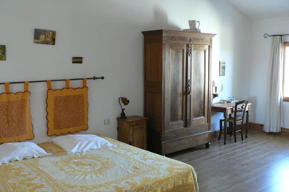 A bedroom with a bed, a wooden wardrobe, a desk with a chair, and a window with curtains.